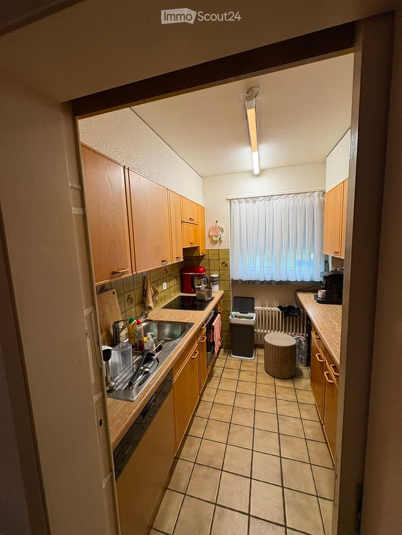 Compact kitchen with wooden cabinets, tiled floor, sink, stove, refrigerator, coffee maker, and a small round stool.