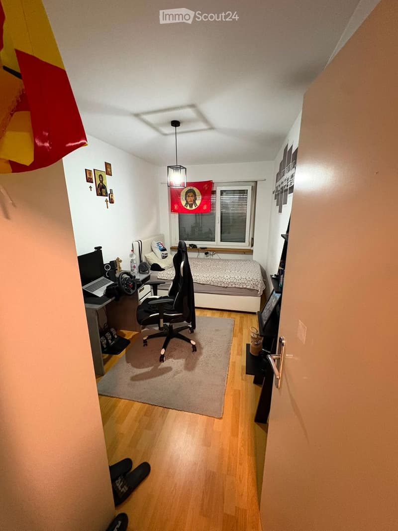 Cozy bedroom with a bed, desk, laptop, gaming chair, and a small rug. The room features wooden flooring and a window with blinds.