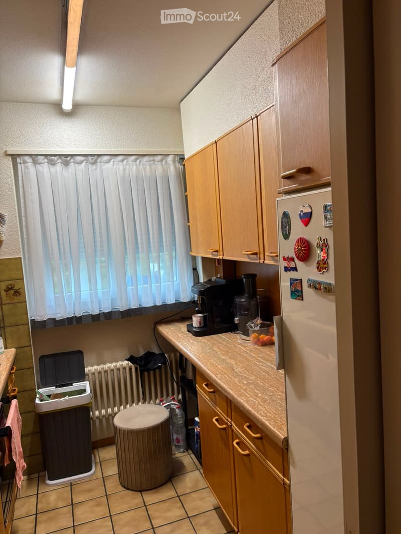 Compact kitchen with wooden cabinets, a refrigerator adorned with magnets, a coffee maker, a food processor, and a small round stool near the trash bin.