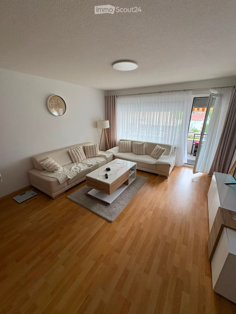 Modern living room with light wooden flooring, a beige sectional sofa, a wooden coffee table, a floor lamp, and a wall-mounted clock.