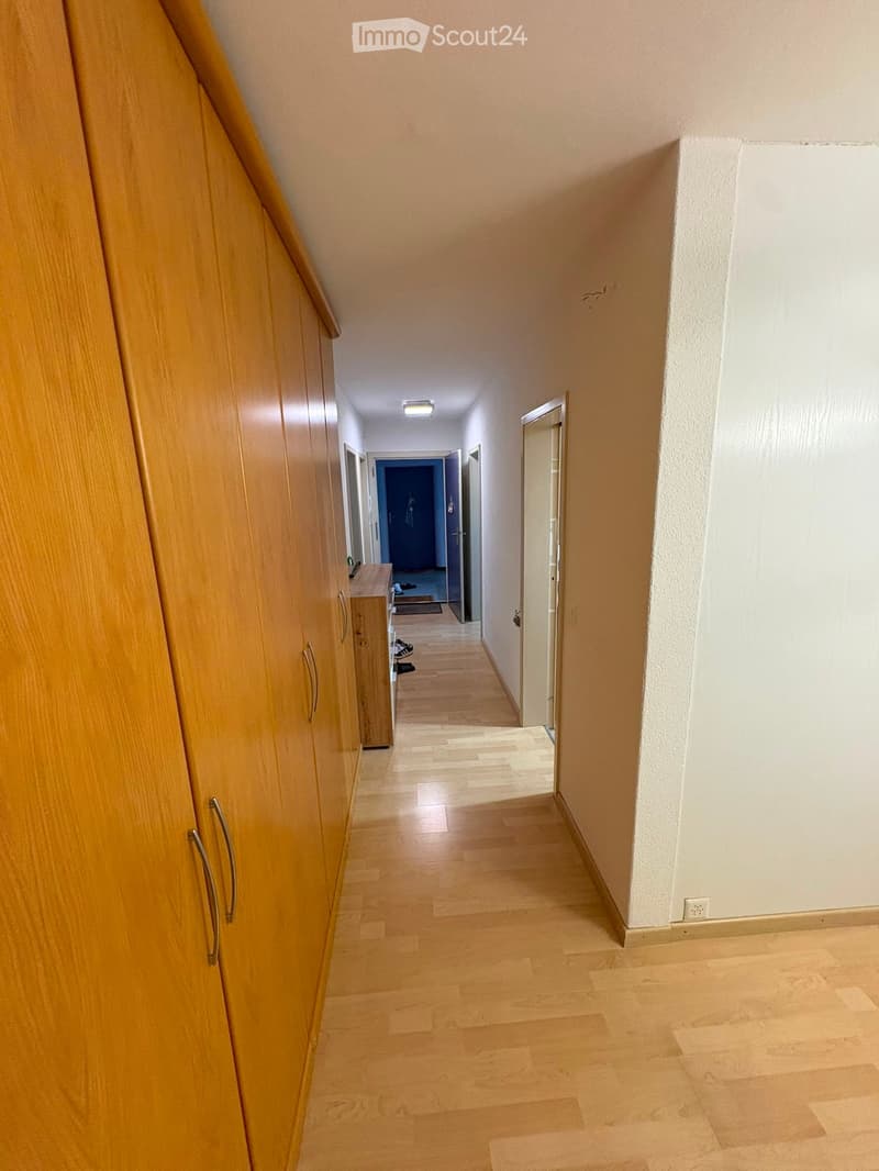 A long, narrow hallway with light wooden flooring, white walls, and a wooden cabinet on the left side. The hallway leads to a blue door at the end and has a small open doorway on the right.