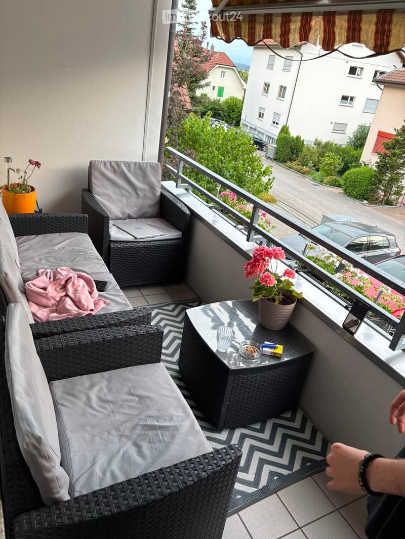 Balcony with wicker furniture, including a sofa, chair, and small table. There are flower pots, a laptop, and a cup on the table. The balcony overlooks a street with houses and greenery.