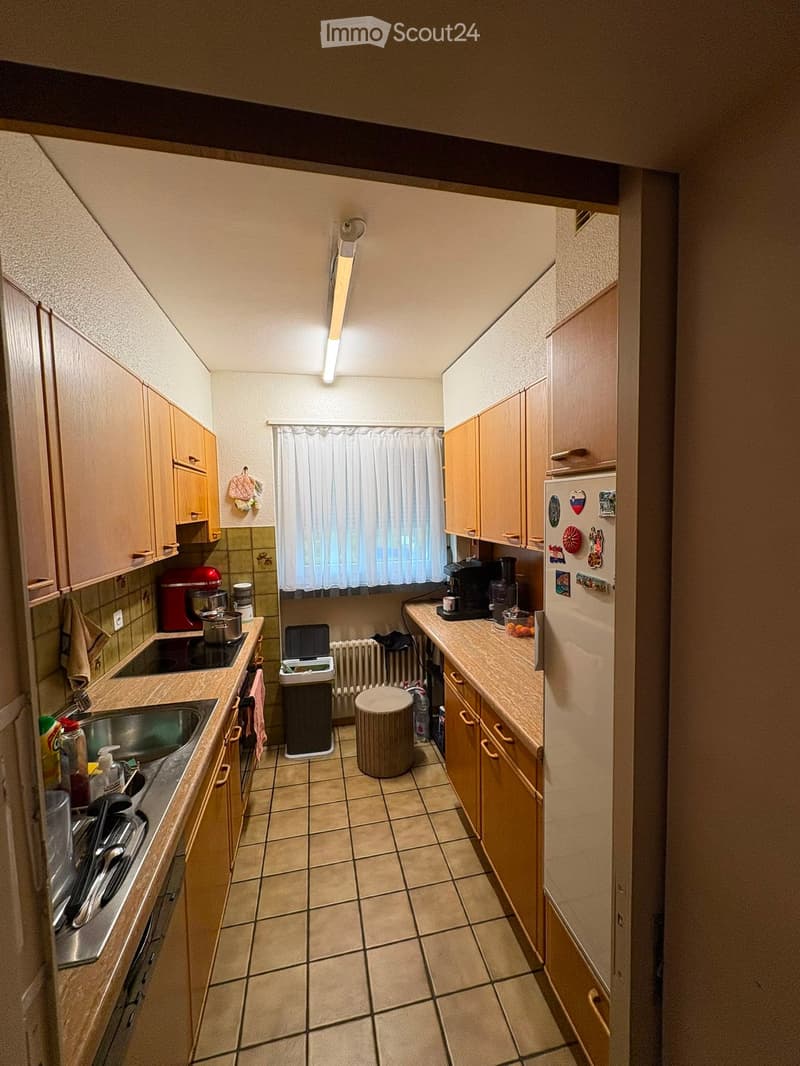 Compact kitchen with light wooden cabinets, tiled floor, and various appliances including a sink, stove, fridge, coffee maker, and blender. A small round stool is placed in the center.