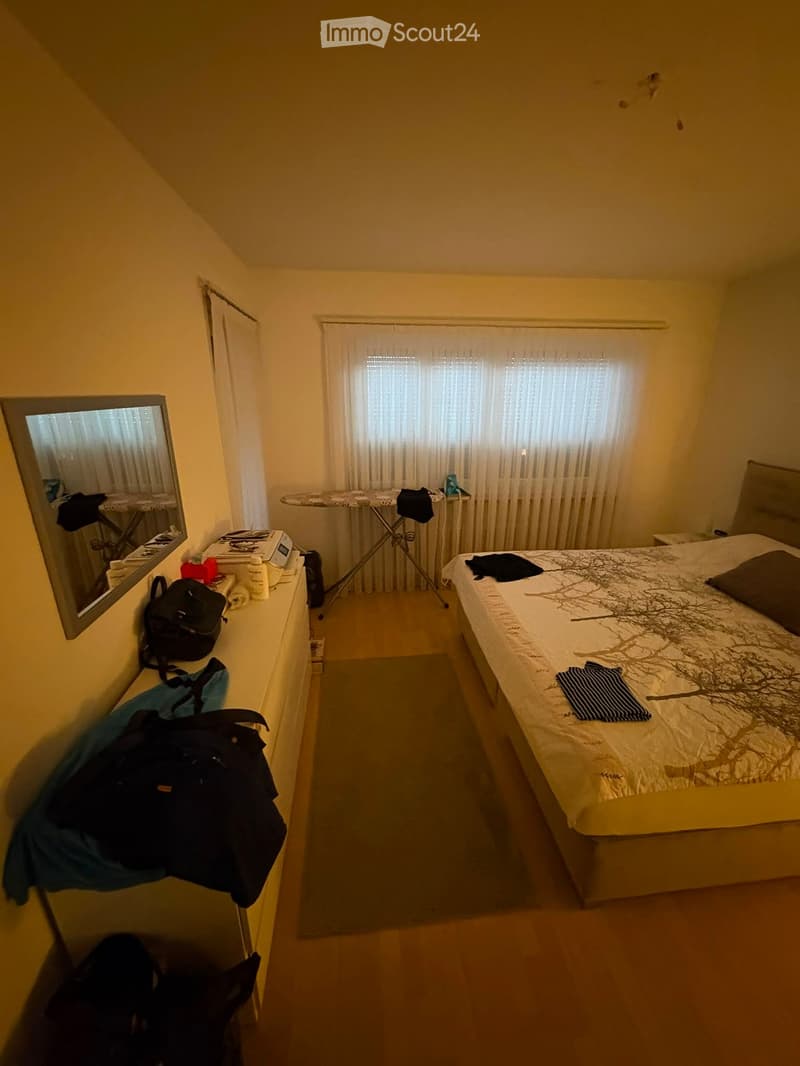Bedroom with a bed, mirror, backpack, ironing board, and nightstand. The room has wooden flooring and large windows with curtains.