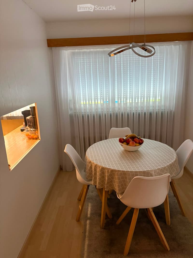 Dining area with a round table covered in a patterned tablecloth, surrounded by four white chairs with wooden legs. A bowl of fruit sits on the table. The room features light-colored walls, a pendant light fixture, and sheer white curtains covering a wind