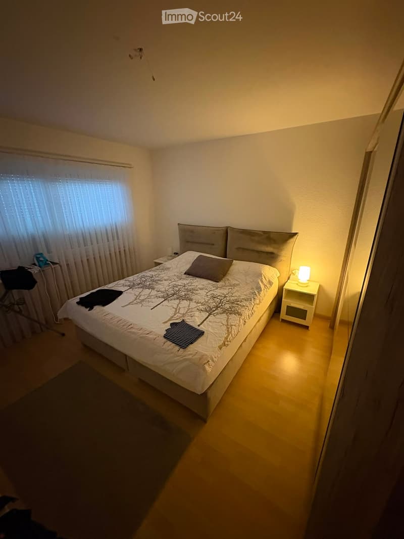 Cozy bedroom with a double bed, nightstand, lamp, and a window with curtains. The bed has a decorative tree-patterned duvet and pillows.