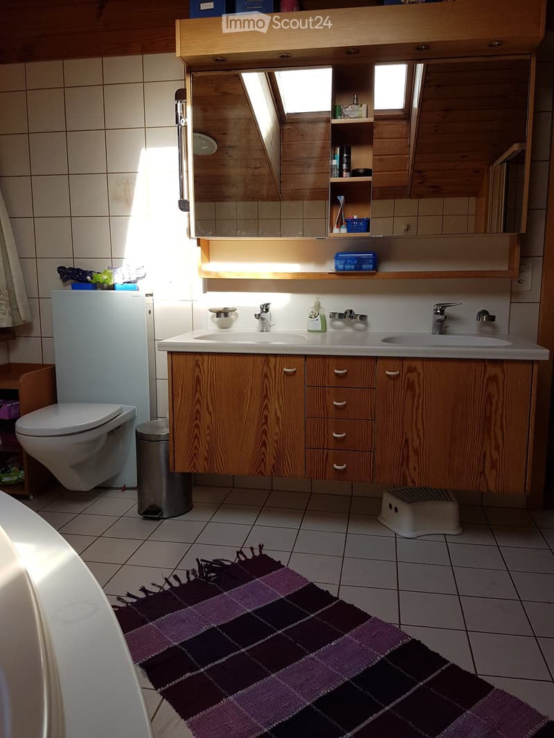 Bathroom with double sink, wooden cabinet, mirror, toilet, and a purple and black checkered rug on the floor.