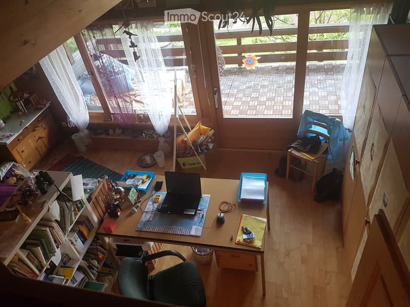 A well-lit room with wooden floors, a large window with white curtains, and a balcony view. The room contains a desk with a laptop, a bookshelf, and a comfortable chair.