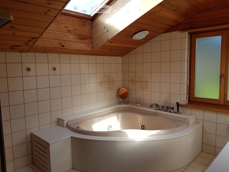 Bathroom with a corner bathtub, tiled walls, wooden ceiling, and frosted window. Features a small mirror and decorative items on the edge of the tub.