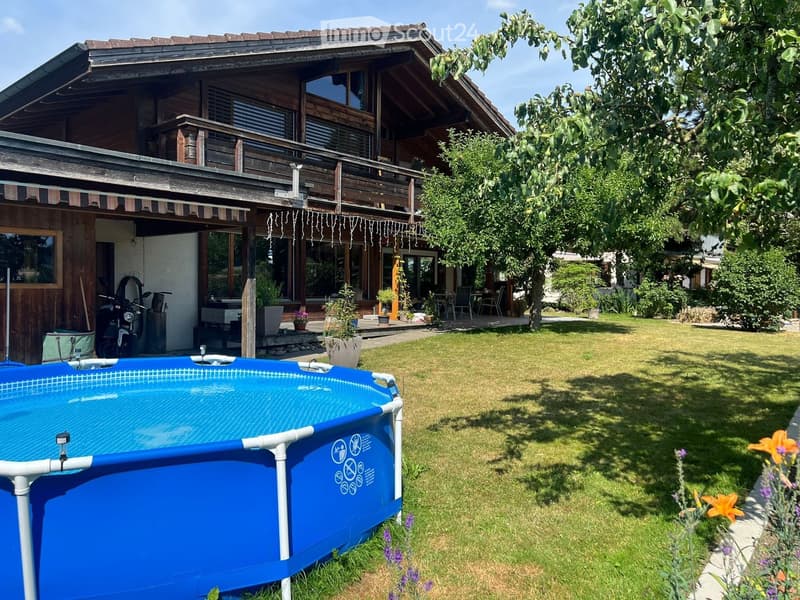 Lush garden with a blue swimming pool, a two-story wooden house with a balcony, and outdoor seating area.