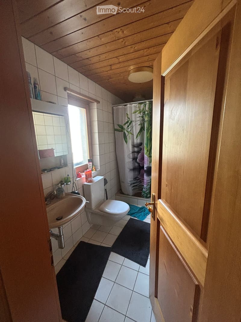 Bathroom with white tiles, a sink, toilet, bathtub with a shower curtain, and a window with a shelf holding toiletries. Wooden ceiling and door frame.