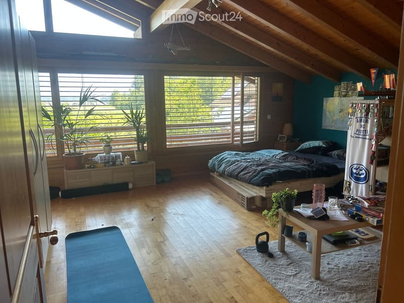 Cozy attic bedroom with wooden slanted ceilings, large windows, a raised platform bed, a small table with items, potted plants, and a yoga mat on the floor.