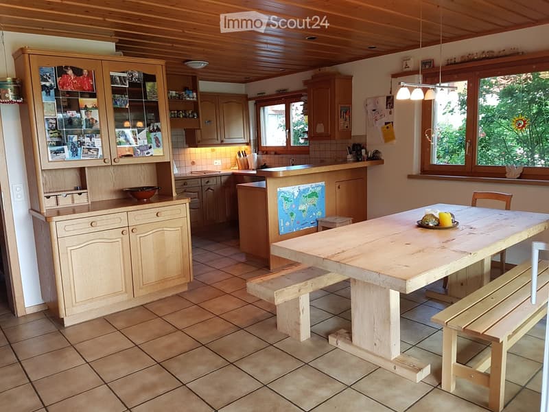 Bright kitchen with wooden cabinets, a large wooden dining table with benches, and a display cabinet with photos and decorative items.