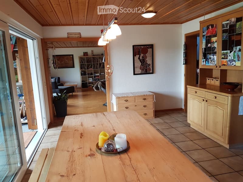 Bright living area with wooden ceiling, tiled floor, and large wooden table with candles. Features a sofa, bookshelf, and cabinet with glass display.