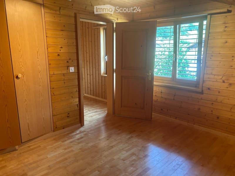 Empty wooden room with light wooden walls, wooden floor, and a door leading to another room. Natural light comes through a window with blinds.