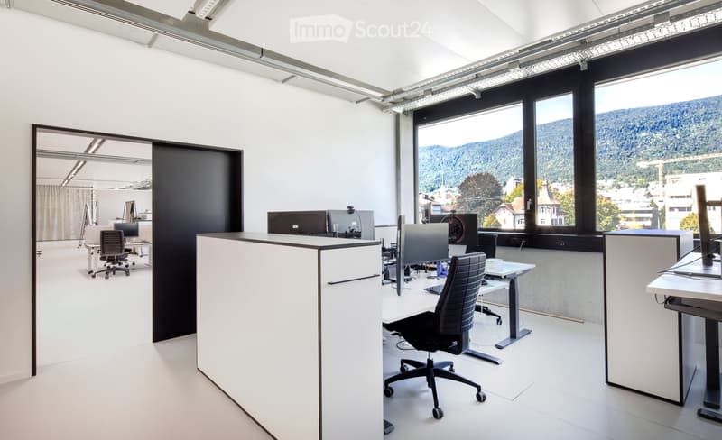 Modern office space with white walls, large windows overlooking a scenic view, and several desks with chairs and computer screens.