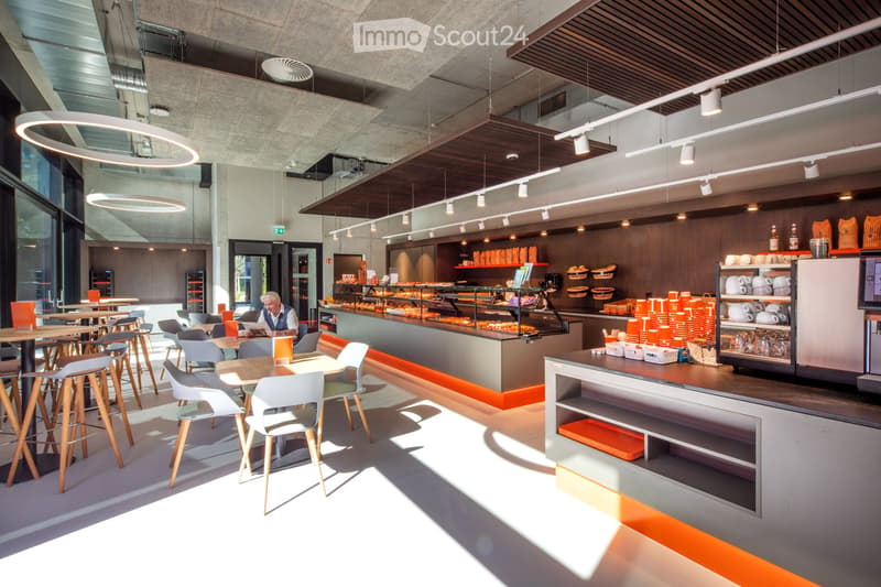 Modern cafeteria with wooden tables, white chairs, and orange stools arranged around a central counter. The counter displays baked goods and beverages, with shelves holding cups and baskets. The space features large windows and pendant lighting.