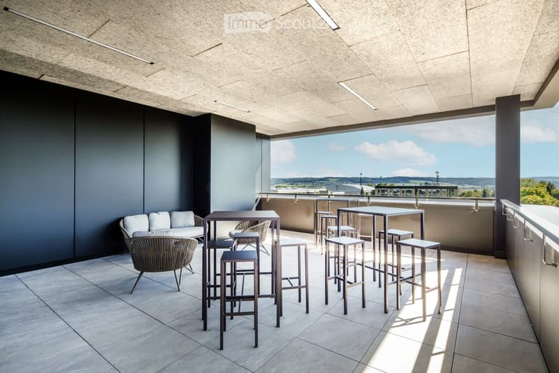 Modern balcony with grey sofa, tables, chairs, and stools, overlooking a scenic landscape.