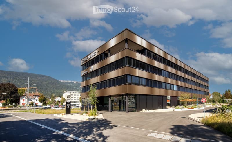 Modern multi-story building with a sleek, contemporary design featuring large glass windows and a metallic facade. The building is labeled "CAMPUS TECHNIK" and is situated in a well-maintained area with surrounding greenery and a clear blue sky.