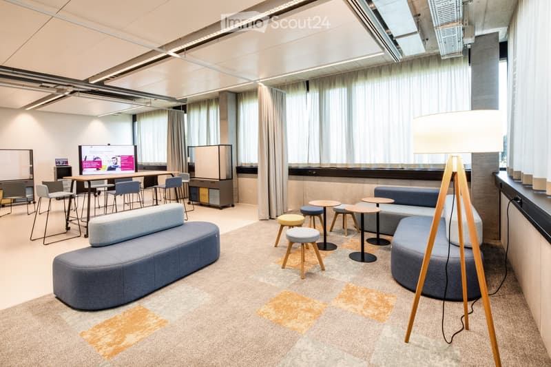 Modern, open-plan lounge area with grey sofas, small tables, stools, and a large screen. Features large windows with curtains and a floor lamp.