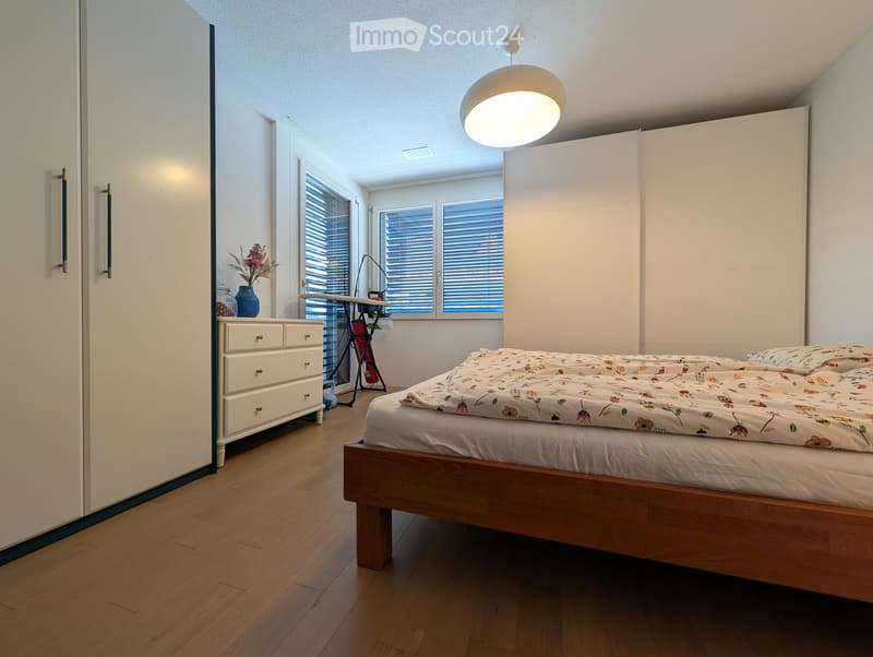 Modern bedroom with wooden flooring, large bed with floral duvet, white dresser with drawers, vase on nightstand, and large windows with blinds.