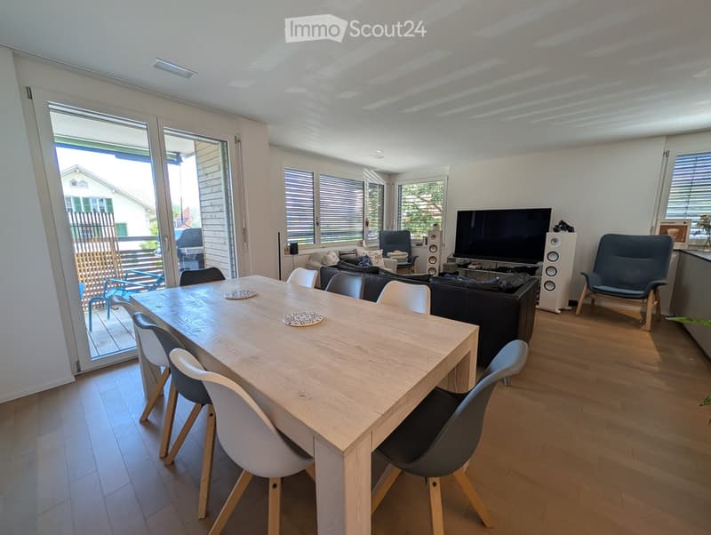 Bright living and dining area with a wooden dining table, modern chairs, a black sofa, and a TV on a stand. Large windows and sliding glass doors lead to a balcony.