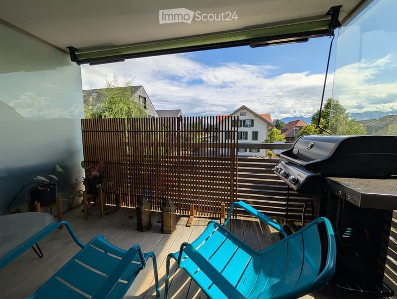 Balcony with wooden decking, two turquoise chairs, a grill, and decorative lanterns, overlooking a residential area with a view of mountains.