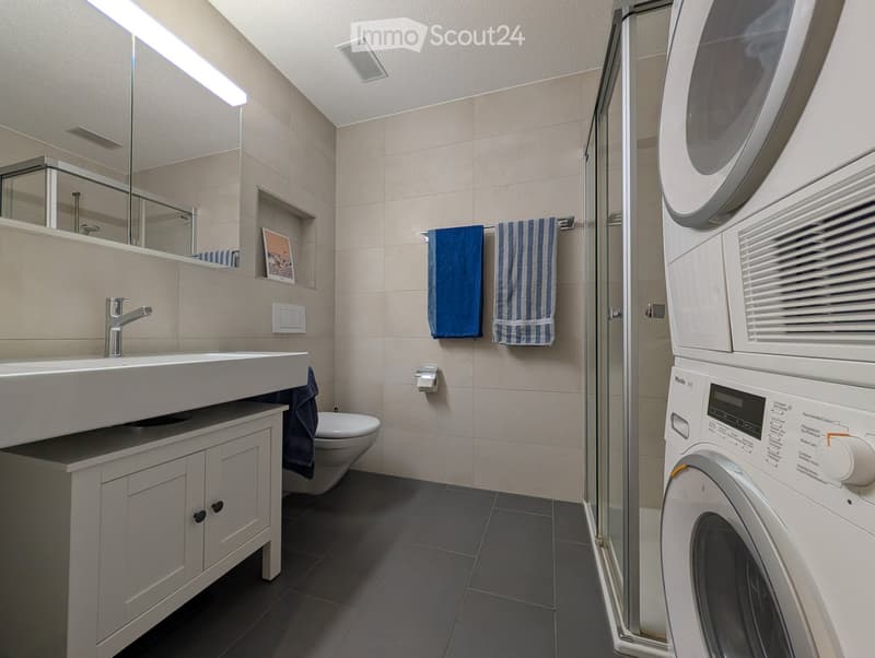 Modern bathroom with a white sink, mirror, cabinet, toilet, and a washing machine. Blue towels are hanging on a rack, and the space features beige tiles and a sleek design.