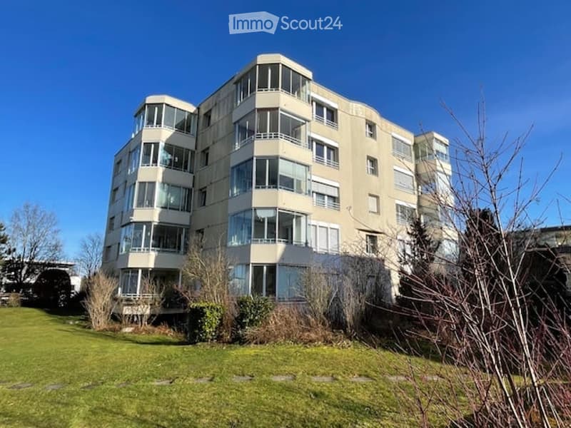 Multi-story building, multiple balconies, glass windows, surrounded by grass and plants
