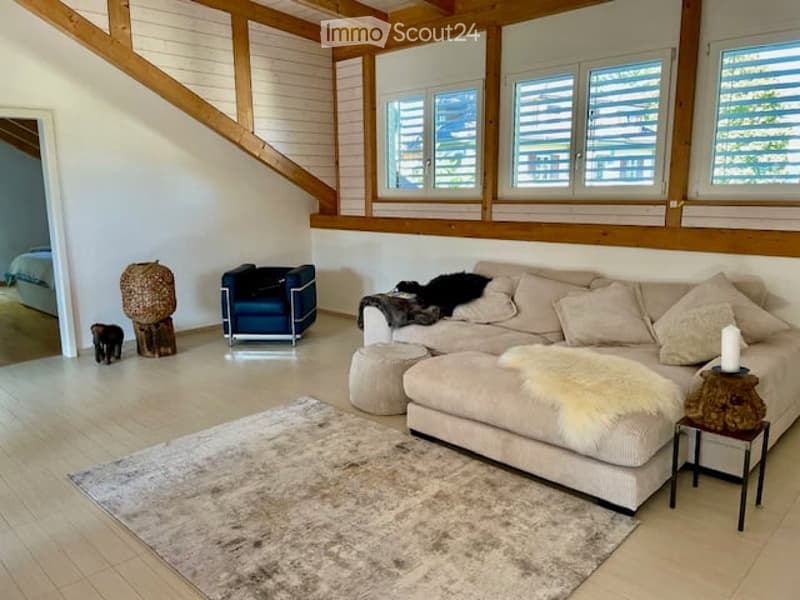Modern living room with large windows, light wood beams, and a cozy seating area featuring a sectional sofa, ottoman, side table, and decorative items.