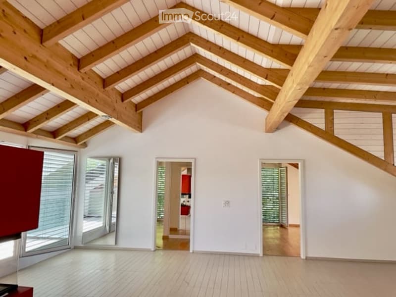 Bright, open loft-style room with exposed wooden beams and white walls. The space features large windows and doors leading to other areas, with a clean, minimalist design and wooden flooring.