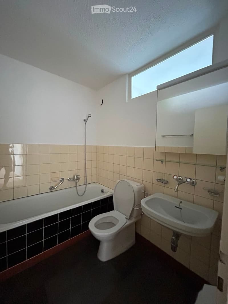 Bathroom with a bathtub, toilet, sink, mirror, and cabinet. The walls are tiled with beige tiles and the floor is dark. There is a window at the top letting in natural light.