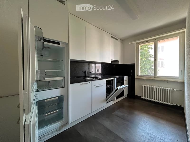 Modern kitchen with white cabinets, stainless steel appliances, and a black countertop. Features a refrigerator, sink, oven, and radiator.