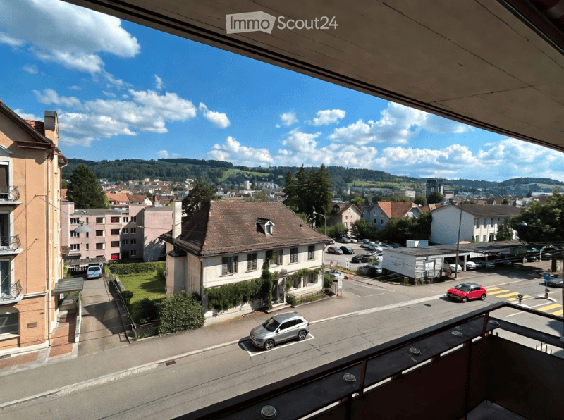 Balcony overlooking a town with houses, parked cars, and a scenic landscape with hills and trees.
