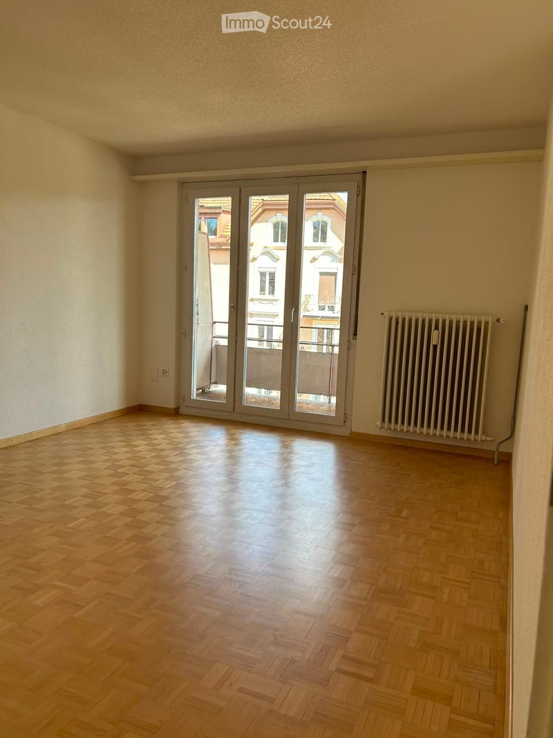 Empty room with wooden herringbone flooring, large sliding glass doors leading to a balcony, and a radiator on the wall.