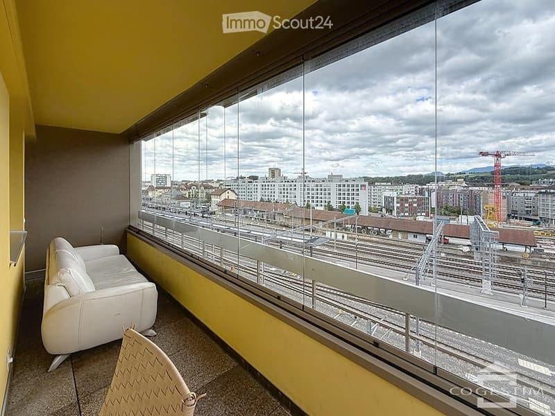 Modern balcony with large glass windows offering a cityscape view, featuring a white leather sofa and a patterned chair.
