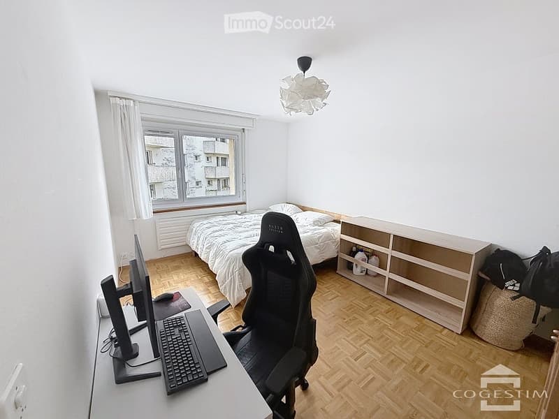 A furnished bedroom with a bed, desk, chair, monitor, keyboard, shelf, and curtain. The room has wooden flooring and large windows.