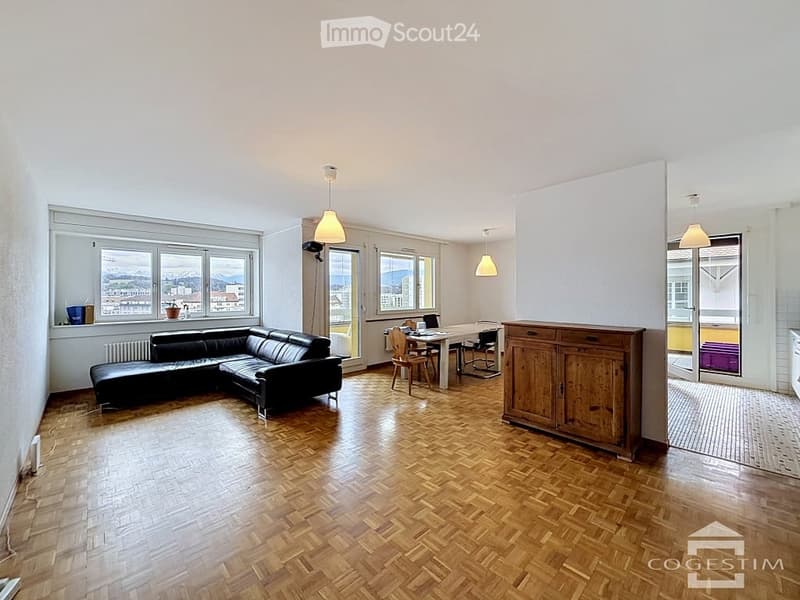 Open-plan living and dining area with wooden flooring, large windows offering a scenic view, a black leather sofa, a dining table with chairs, and a wooden cabinet.