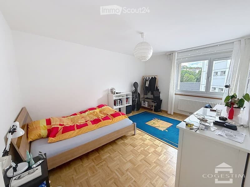 Bright bedroom with wooden flooring, large window with white curtains, bed with colorful quilt, nightstand, desk with chair, bookshelf, guitar, coat rack, and a small plant on the desk.