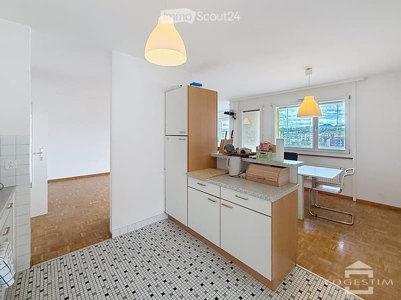 Modern kitchen with white cabinets, granite countertop, refrigerator, and a small dining area with a table and chairs. Large windows offer a scenic view.