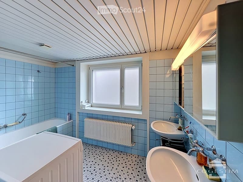 Bathroom with blue tiles, white radiator, double sink, bathtub, and a window providing natural light.