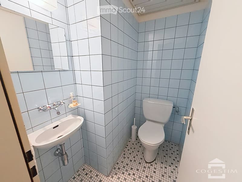 A small bathroom with light blue tiled walls and a black and white checkered floor. It features a white toilet, a white sink with a chrome faucet, and a small mirror above the sink. A toilet brush is placed next to the toilet.
