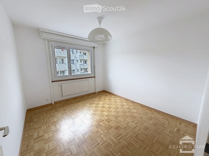 Empty room with wooden parquet flooring, white walls, large window with radiator below, and a modern pendant light fixture.