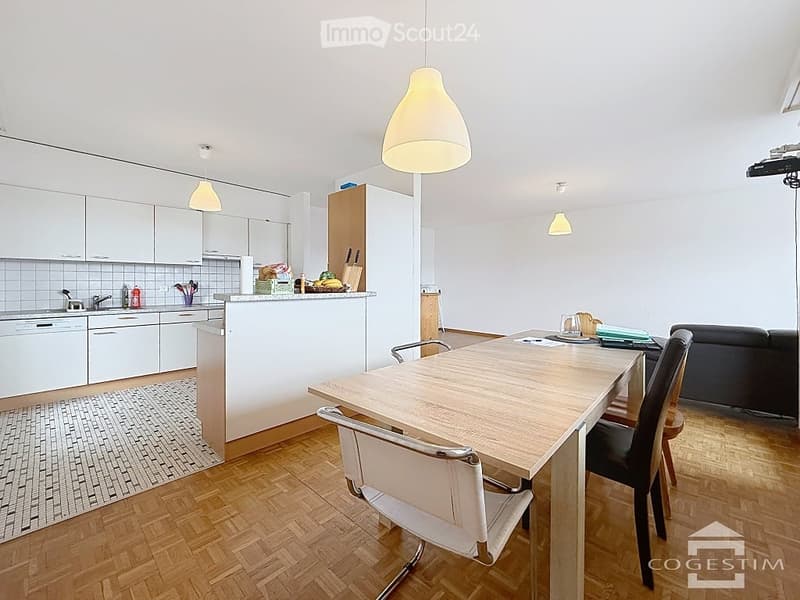 Open-plan living and dining area with a modern kitchen, white cabinets, a central island, and a dining table with chairs. The space features wooden flooring, pendant lights, and a sleek sofa.