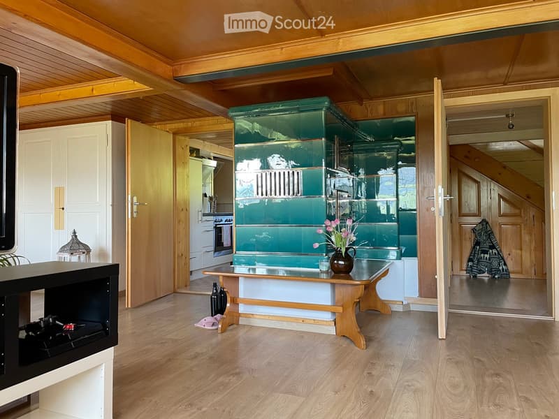 A cozy, well-lit room with wooden floors and a slanted wooden ceiling. Features include a green stove, a small wooden table with a vase of flowers, a bench, and a doorway leading to another room.