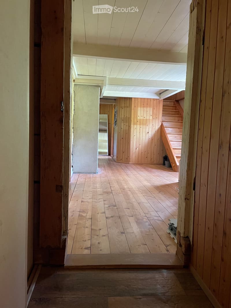 Wooden-floored hallway with exposed wooden beams and a staircase leading upwards, featuring wooden panel walls and a partially open ceiling with skylight.