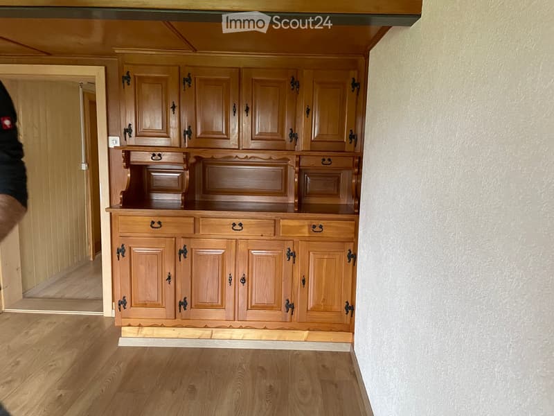 A wooden cabinet with multiple doors and drawers is installed against a wall in a room with wooden flooring and light-colored walls.