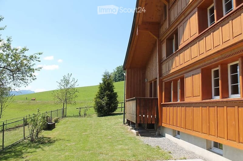 A two-story wooden building with white-framed windows and balconies, situated in a grassy field with trees and a clear blue sky in the background.