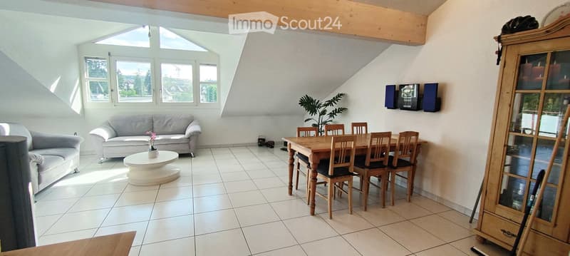 Bright living and dining area with white tiled flooring, large windows, grey sofa, coffee table, dining table with chairs, and a wooden cabinet.