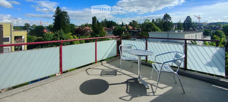 Balcony with a round metal table and two matching chairs, surrounded by a glass railing and overlooking a green, tree-filled area with houses in the background.